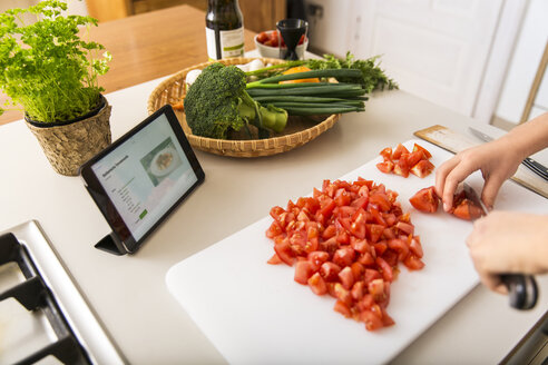 Young woman cutting tomatoes on kitchen counter and reading recipe on tablet - JHAF00026