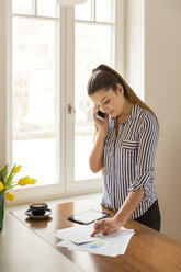 Junge Frau am Telefon bei der Arbeit an gedruckten Tabellen auf einem Tresen zu Hause - JHAF00029