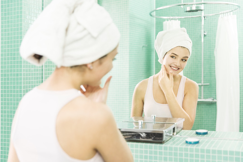 Young woman applying facial moisturizer at mirror in bathroom stock photo