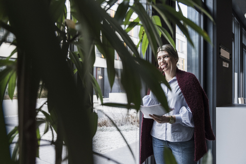 Lächelnde junge Frau am Fenster stehend mit Handy und Dokumenten, lizenzfreies Stockfoto