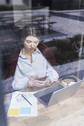 Young woman with cell phone, laptop and documents behind windowpane - UUF12835