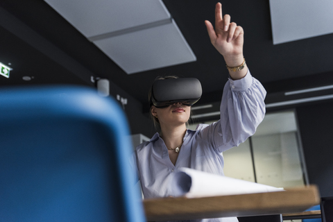 Junge Frau mit VR-Brille am Tisch im Büro, lizenzfreies Stockfoto