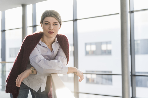 Portrait of confident young woman leaning on railing in bright office building stock photo