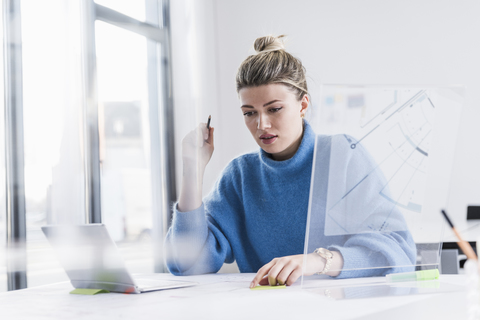 Junge Frau mit Laptop und transparentem Entwurf arbeitet an einem Plan am Schreibtisch im Büro, lizenzfreies Stockfoto