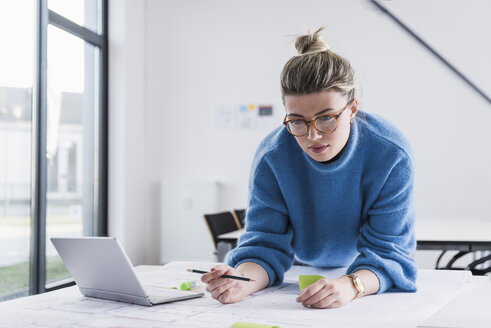 Junge Frau mit Laptop arbeitet an einem Plan am Schreibtisch im Büro - UUF12856