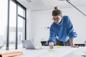 Junge Frau mit Laptop arbeitet an einem Plan am Schreibtisch im Büro - UUF12857