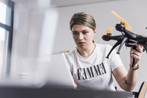 Junge Frau mit Laptop am Schreibtisch, die eine Drohne hält, lizenzfreies Stockfoto