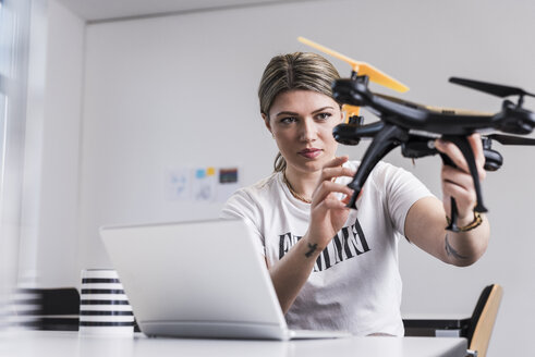 Junge Frau mit Laptop am Schreibtisch, die eine Drohne hält - UUF12862