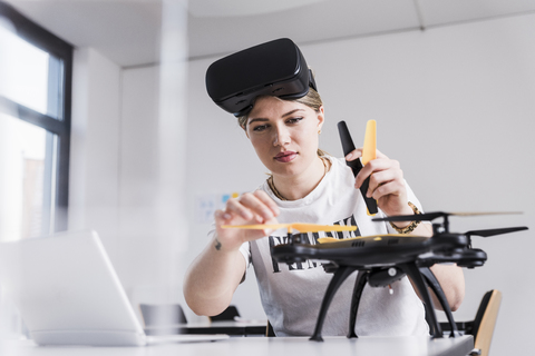 Junge Frau mit Laptop und VR-Brille am Schreibtisch bei der Untersuchung einer Drohne, lizenzfreies Stockfoto