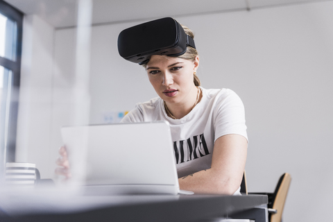 Junge Frau mit Laptop und VR-Brille arbeitet am Schreibtisch im Büro, lizenzfreies Stockfoto