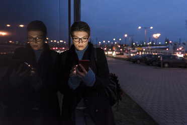 Junge Frau lehnt nachts an einer Glasfront und benutzt ein Mobiltelefon - UUF12867