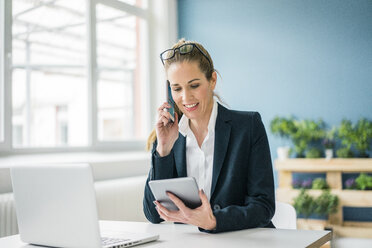 Geschäftsfrau sitzt am Schreibtisch, telefoniert und schaut auf ein digitales Tablet - MOEF00884