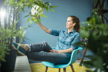 Beautiful woman sitting in her home, decorated with plants, looking at globe - MOEF00888