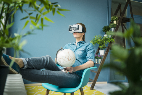 Beautiful woman sitting in her home, decorated with plants, holding globe, wearing VR glasses stock photo