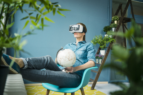 Schöne Frau sitzt in ihrem Haus, dekoriert mit Pflanzen, hält Globus, trägt VR-Brille, lizenzfreies Stockfoto