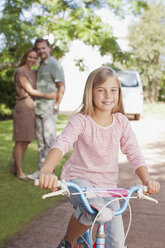 Porträt eines lächelnden Mädchens auf einem Fahrrad mit Eltern im Hintergrund - CAIF00040