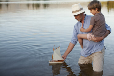 Grandfather and grandson wading in lake with toy sailboat - CAIF00090
