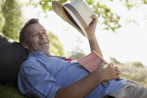 Porträt eines lächelnden Mannes, der mit Buch und Hut im Gras liegt, lizenzfreies Stockfoto