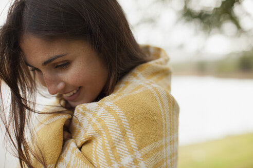 Nahaufnahme einer lächelnden, in eine Decke gehüllten Frau am Seeufer - CAIF00125