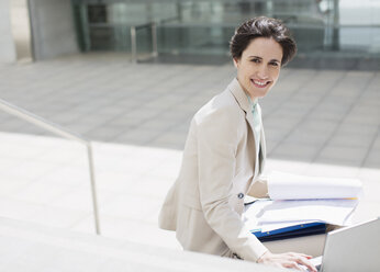 Portrait of smiling businesswoman with laptop and paperwork outside building - CAIF00272