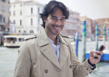 Porträt eines lächelnden Mannes mit Handy in der Hand am Hafen von Venedig - CAIF00478