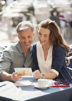 Lächelndes Paar betrachtet eine Postkarte in einem Straßencafé - CAIF00502