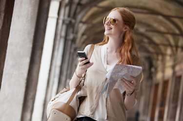 Smiling woman holding cell phone and map in corridor - CAIF00517