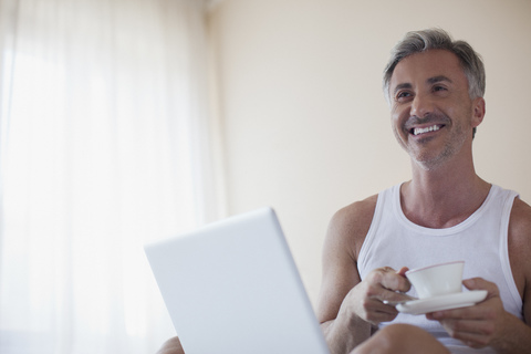 Lächelnder Mann trinkt Kaffee und benutzt Laptop im Schlafzimmer, lizenzfreies Stockfoto