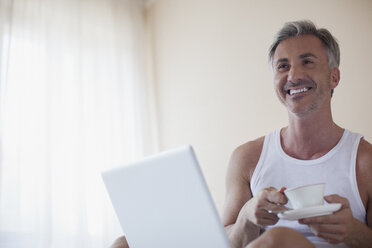 Smiling man drinking coffee and using laptop in bedroom - CAIF00519