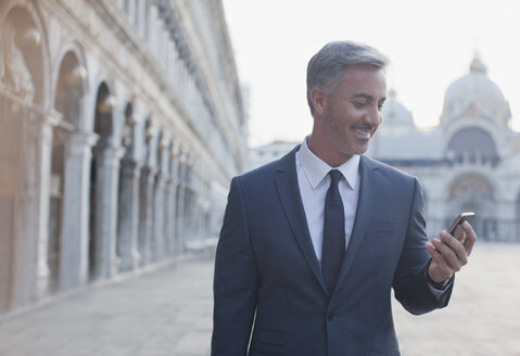 Lächelnder Geschäftsmann überprüft sein Handy auf dem Markusplatz in Venedig - CAIF00550