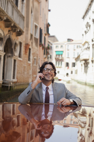 Lächelnder Geschäftsmann, der mit seinem Handy telefoniert und mit einem Boot durch einen Kanal in Venedig fährt, lizenzfreies Stockfoto