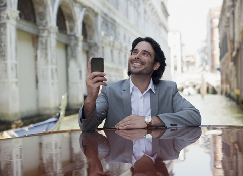 Lächelnder Geschäftsmann mit Handy in der Hand und an ein Boot im Kanal in Venedig gelehnt, lizenzfreies Stockfoto