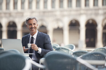 Lächelnder Geschäftsmann trinkt Espresso und benutzt einen Laptop in einem Straßencafé - CAIF00572