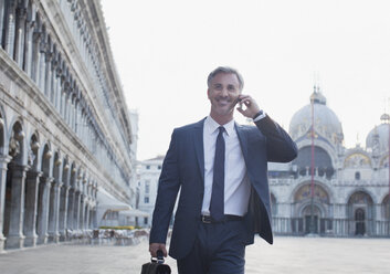 Lächelnder Geschäftsmann, der mit seinem Handy telefoniert und über den Markusplatz in Venedig läuft - CAIF00576