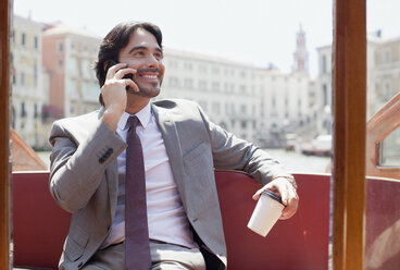 Smiling businessman with coffee talking on cell phone on boat in Venice - CAIF00590