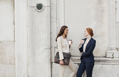 Lächelnde Geschäftsfrauen, die Kaffee trinken und sich an einer Hauswand unterhalten - CAIF00595