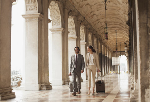 Lächelnder Geschäftsmann und Geschäftsfrau, die Koffer durch einen Korridor in Venedig ziehen - CAIF00623