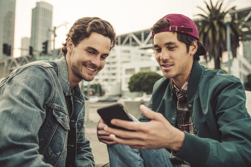 Two smiling young men sharing cell phone outdoors - SUF00536