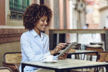 Lächelnde Frau mit Afrofrisur sitzt in einem Café im Freien und benutzt ein Tablet - JSMF00013