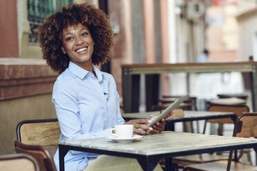 Lächelnde Frau mit Afrofrisur sitzt im Café im Freien mit Tablet - JSMF00014