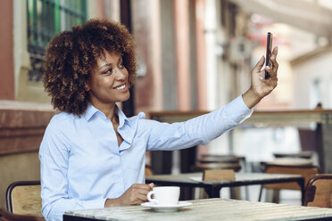 Smiling woman with afro hairstyle sitting in outdoor cafe taking a selfie - JSMF00015