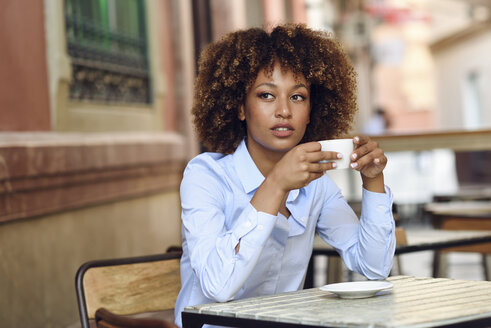 Frau mit Afrofrisur sitzt in einem Café im Freien und trinkt Kaffee - JSMF00019
