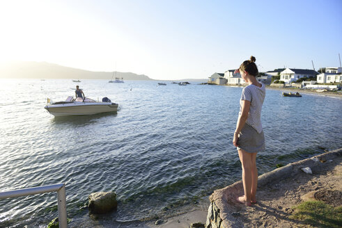 Junge Frau steht an der Küste und betrachtet einen Mann auf einem Boot im Meer - ECPF00201