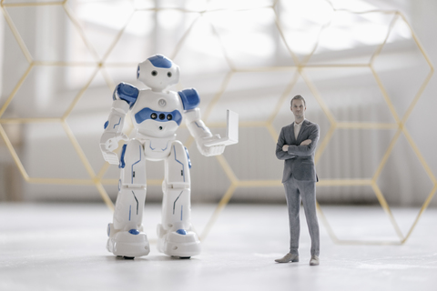 Miniatur-Geschäftsmann-Figur neben Roboter mit Laptop stehend, lizenzfreies Stockfoto
