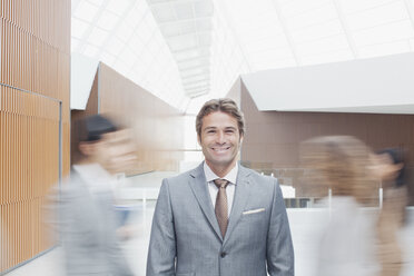 Porträt eines selbstbewussten Geschäftsmannes mit vorbeieilenden Mitarbeitern in der Lobby - CAIF01316