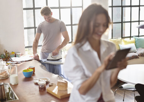 Frau benutzt digitales Tablet, während ihr Freund in der Küche bügelt - CAIF01548