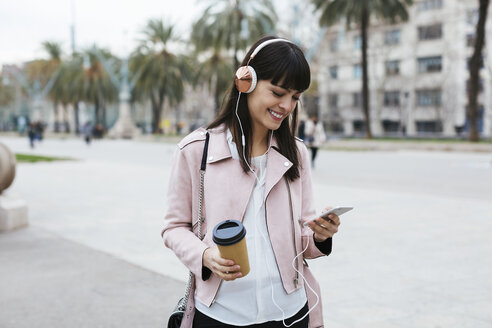 Spanien, Barcelona, lächelnde Frau mit Kaffee, Mobiltelefon und Kopfhörer in der Stadt - EBSF02152