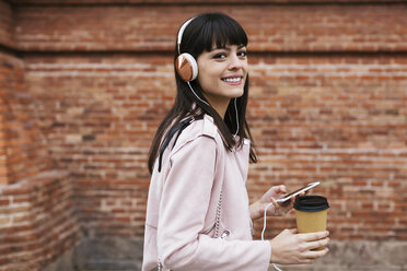 Porträt einer lächelnden Frau mit Kaffee, Mobiltelefon und Kopfhörern an einer Backsteinmauer - EBSF02154