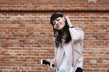 Smiling woman with cell phone listening to music on headphones at brick wall - EBSF02157