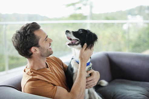 Lächelnder Mann streichelt Hund auf Sofa - CAIF02215